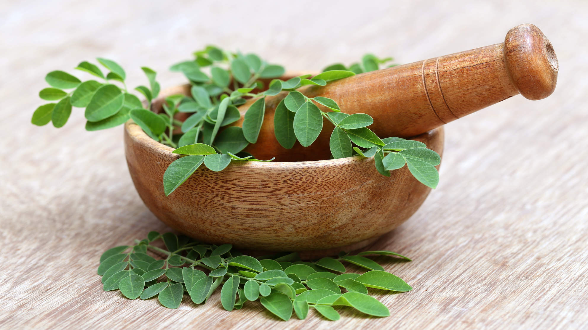Fresh green moringa leaves piled around a rustic wooden mortar and pestle on a wooden surface, earthy and natural tones